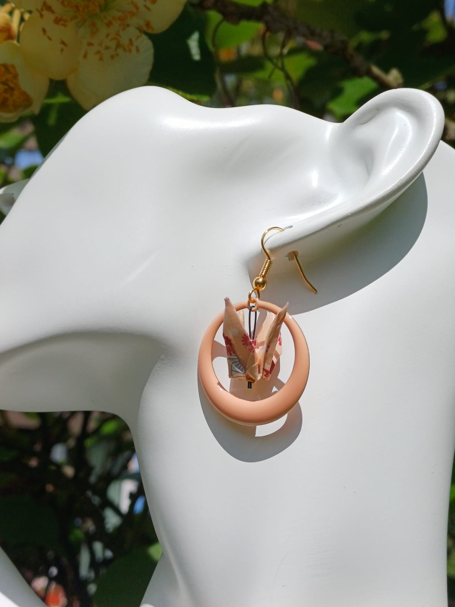 Boucles d'oreilles origami grue beige dans créole résine rose poudré, portées sur mannequin bijou fond végétal