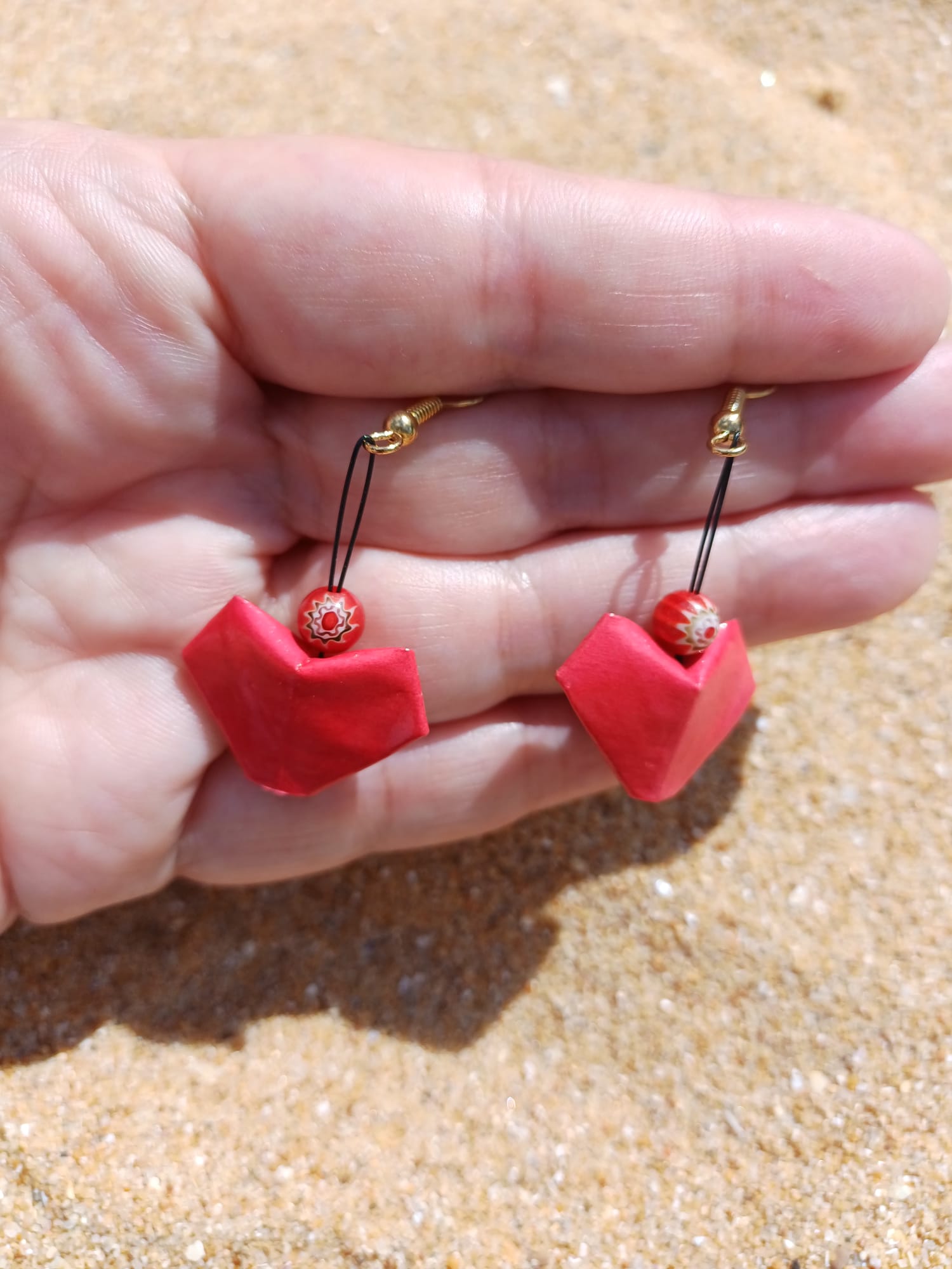 Boucles d'oreilles origami cœur rouge faites main tenues dans une main, fond sable