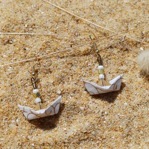 Boucles d'oreilles origami petit bateau blanc et or posées sur sable doré avec herbes sèches