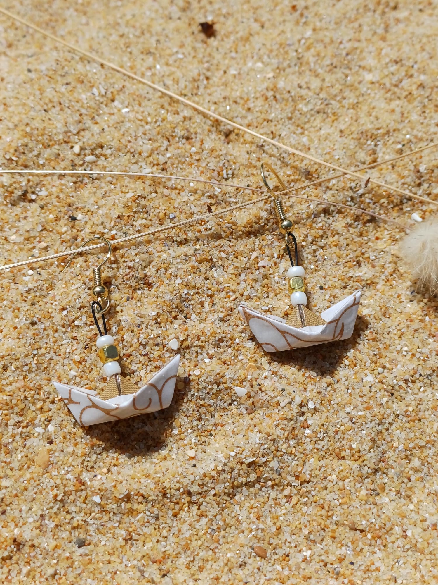 Boucles d'oreilles origami petit bateau blanc et or posées sur sable doré avec herbes sèches
