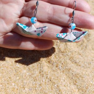 Paire de boucles d'oreilles origami bateau fleuri turquoise et rose tenues dans une main ouverte, fond sable