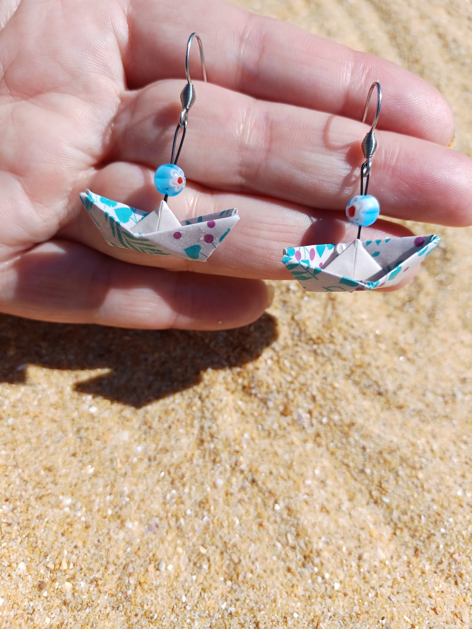 Paire de boucles d'oreilles origami bateau fleuri turquoise et rose tenues dans une main ouverte, fond sable