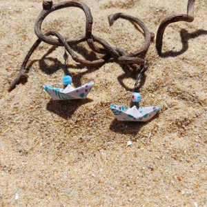 Boucles d'oreilles origami bateau fleuri posées sur sable doré avec branches de bois flotté