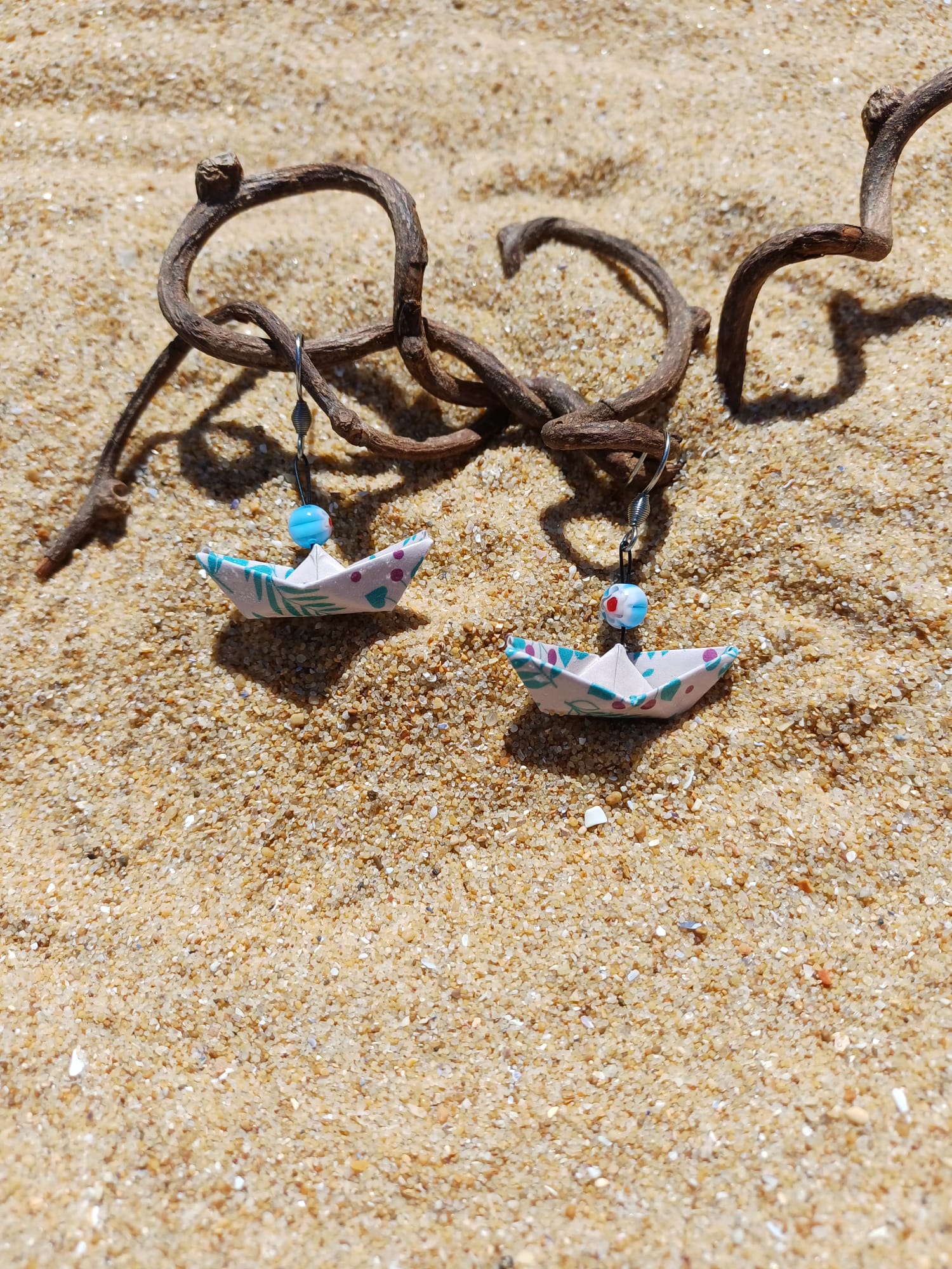 Boucles d'oreilles origami bateau fleuri posées sur sable doré avec branches de bois flotté