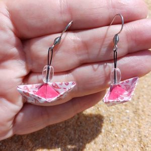 Boucles d'oreilles origami bateau rose paisley avec intérieur fuchsia et perle transparente tenues dans une main ouverte fond sable