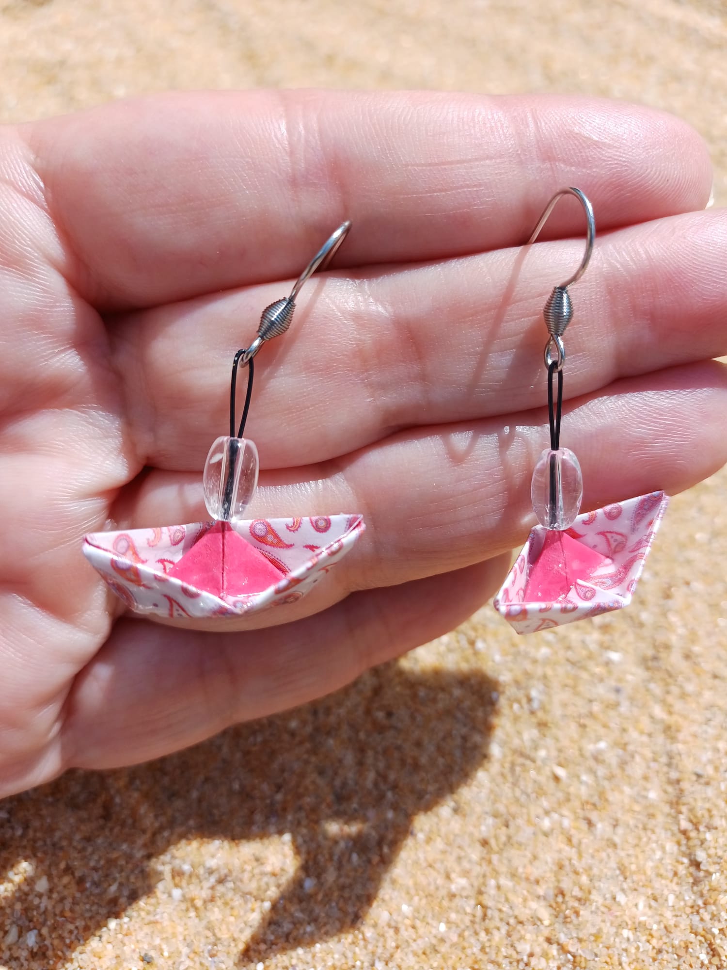 Boucles d'oreilles origami bateau rose paisley avec intérieur fuchsia et perle transparente tenues dans une main ouverte fond sable