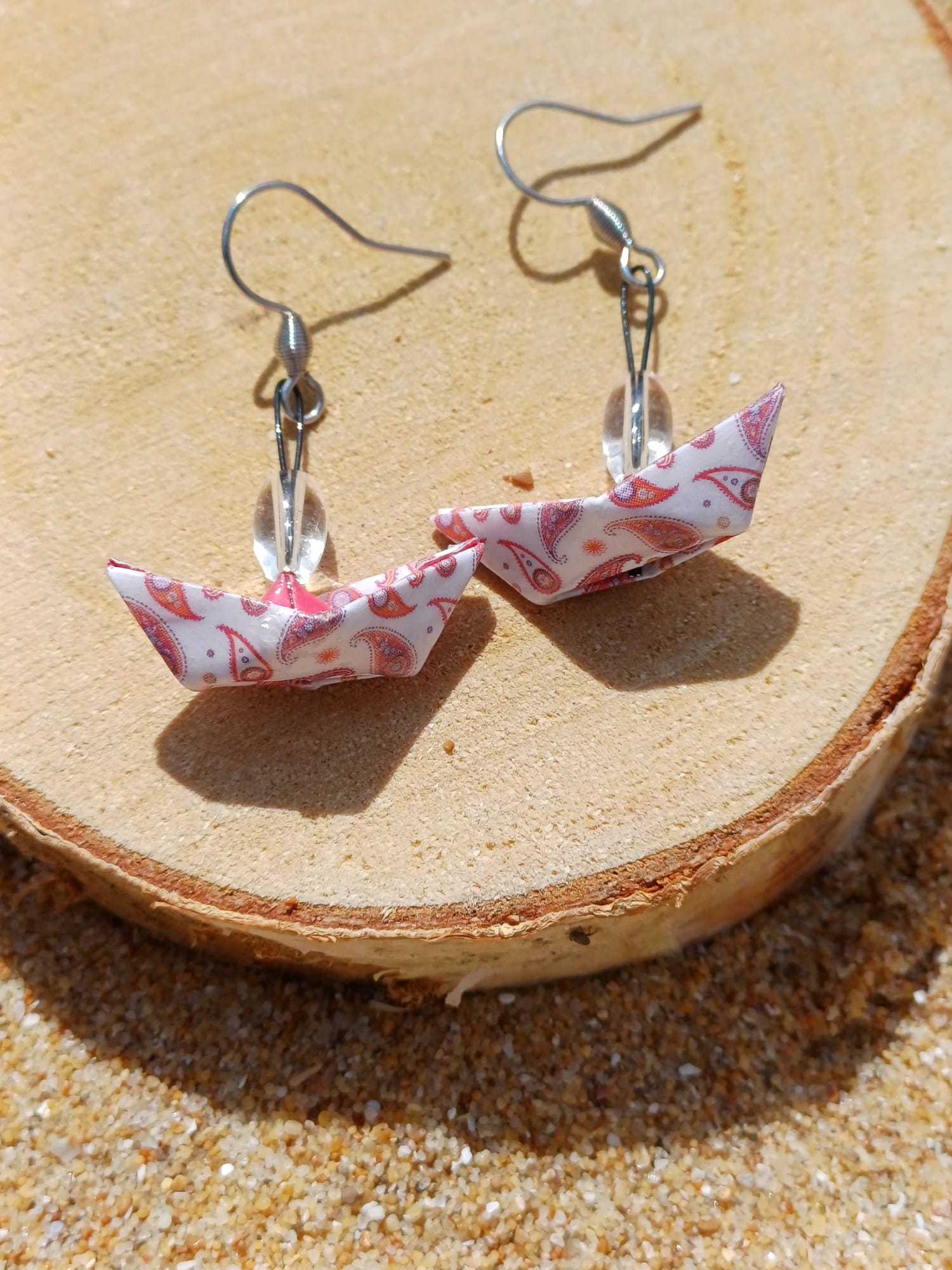 Paire de boucles d'oreilles origami bateau blanc motifs paisley rose posées sur rondelle de bois dans le sable