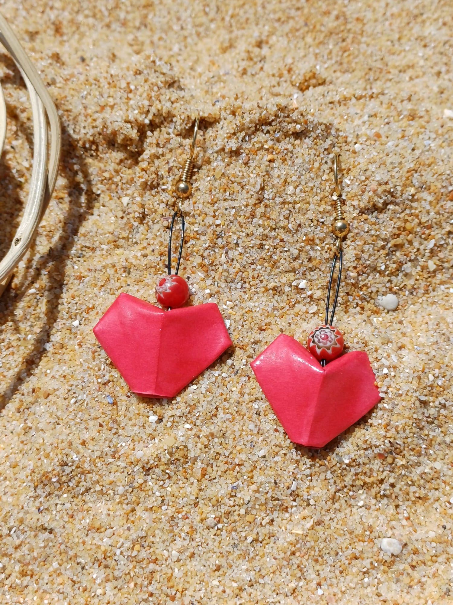 Paire de boucles d'oreilles origami en forme de cœur rouge nacré avec perles millefiori, posées sur le sable