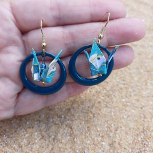 Paire de boucles d'oreilles origami grue blanche créole résine bleu canard tenues dans une main ouverte, fond sable
