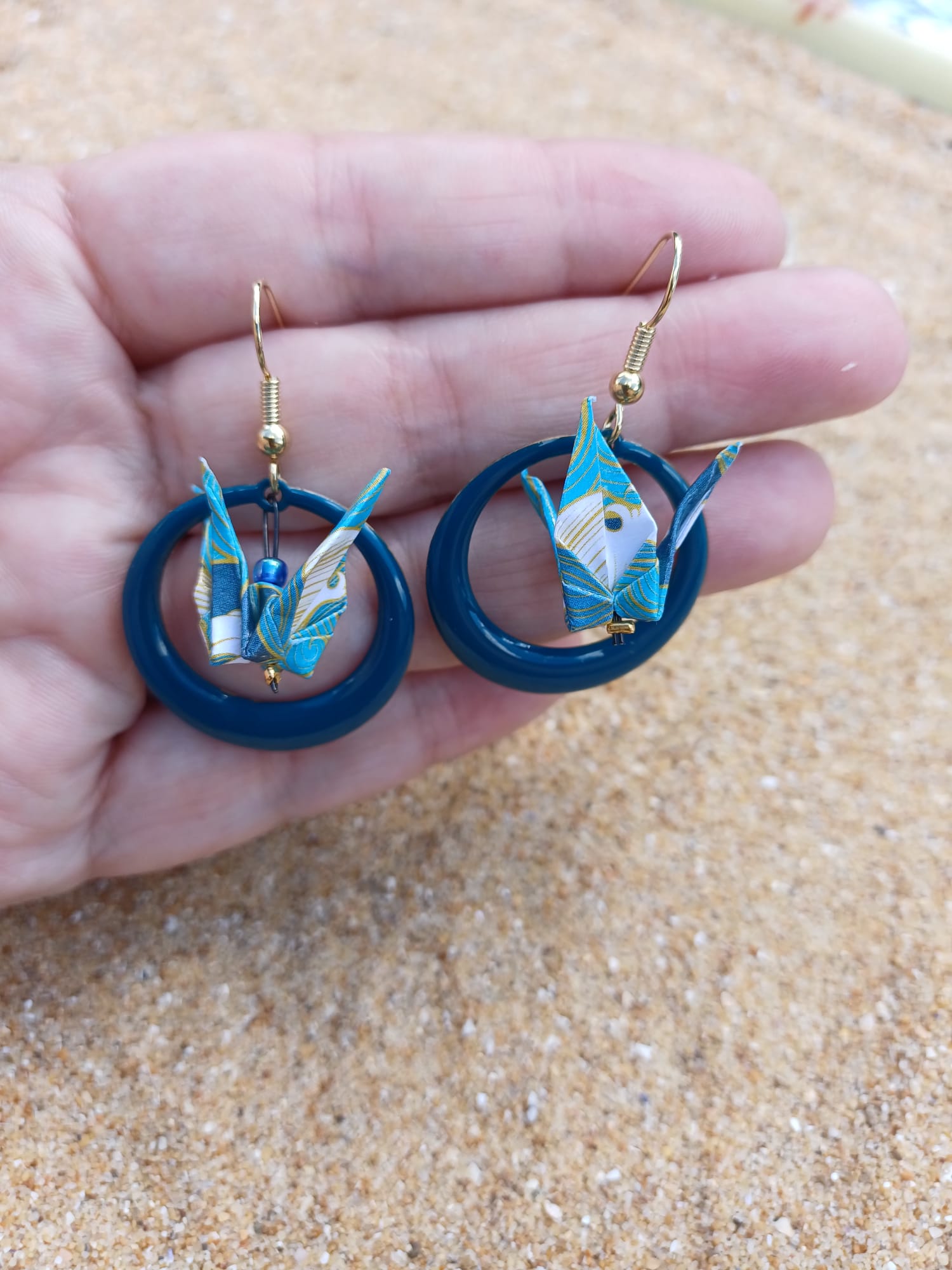Paire de boucles d'oreilles origami grue blanche créole résine bleu canard tenues dans une main ouverte, fond sable