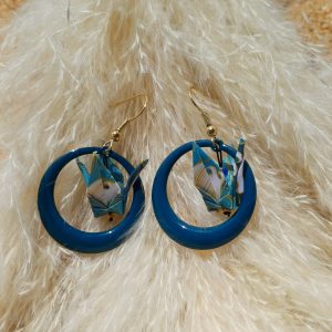 Paire de boucles d'oreilles origami grue bleu canard et plumes dorées posées sur herbes de pampa blanches fond sable