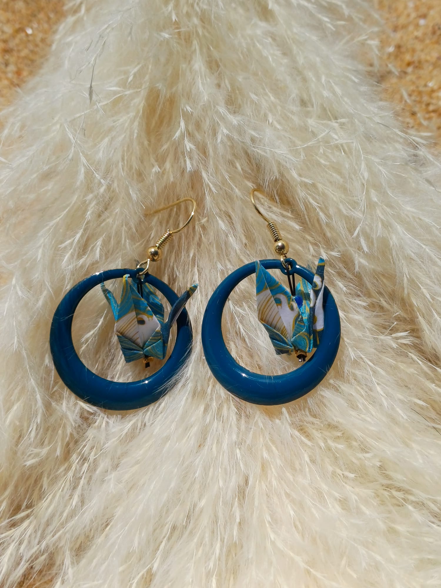 Paire de boucles d'oreilles origami grue bleu canard et plumes dorées posées sur herbes de pampa blanches fond sable