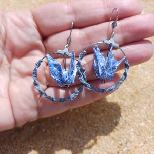 Paire de boucles d'oreilles origami grue bleue dans créole macramé tenues dans une main ouverte, fond sable