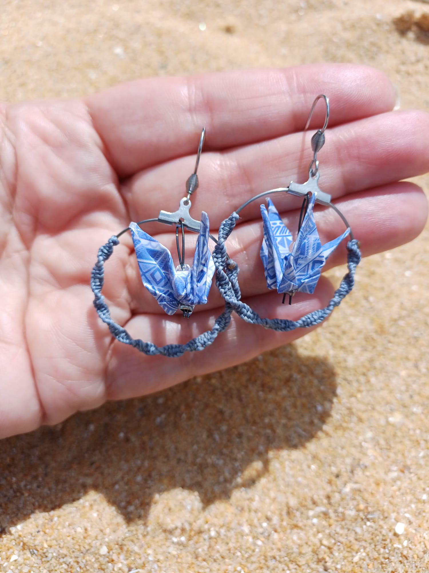 Paire de boucles d'oreilles origami grue bleue dans créole macramé tenues dans une main ouverte, fond sable