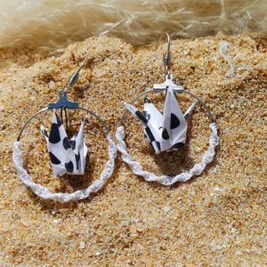 Paire de boucles d'oreilles origami grue blanc à taches noires dans créole macramé blanc, posées dans le sable avec herbes de pampa