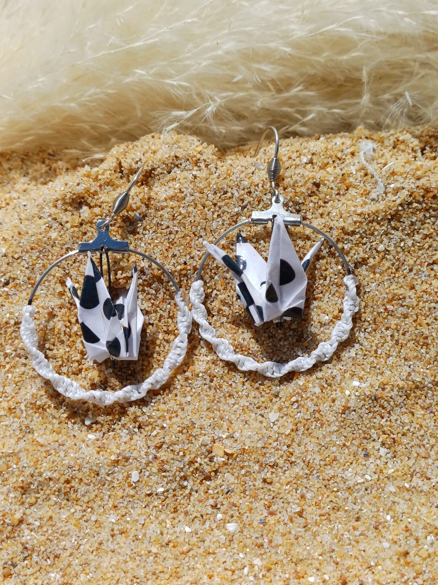 Paire de boucles d'oreilles origami grue blanc à taches noires dans créole macramé blanc, posées dans le sable avec herbes de pampa