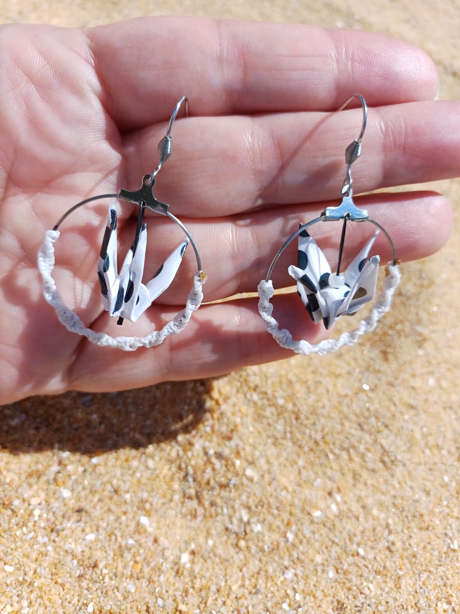 Paire de boucles d'oreilles origami grue blanc taches noires créole macramé blanc tenues dans une main ouverte fond sable