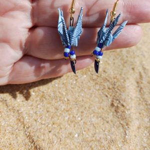 Paire de boucles d'oreilles origami grue bleu ciel avec sequin éventail bleu cobalt et perles rocaille, tenues dans une main ouverte fond sable