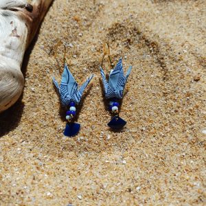 Boucles d'oreilles origami grue bleu ciel et sequin éventail émaillé posées dans le sable avec bois flotté