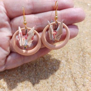 Paire de boucles d'oreilles origami grue dans créole résine rose poudré tenues dans une main, fond sable