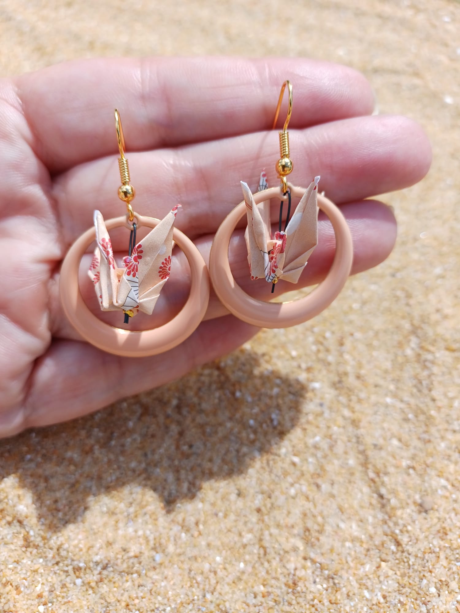 Paire de boucles d'oreilles origami grue dans créole résine rose poudré tenues dans une main, fond sable