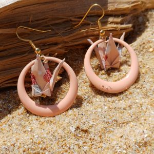 Boucles d'oreilles origami grue beige à motifs floraux dans créole rose poudré, posées sur sable avec bois flotté