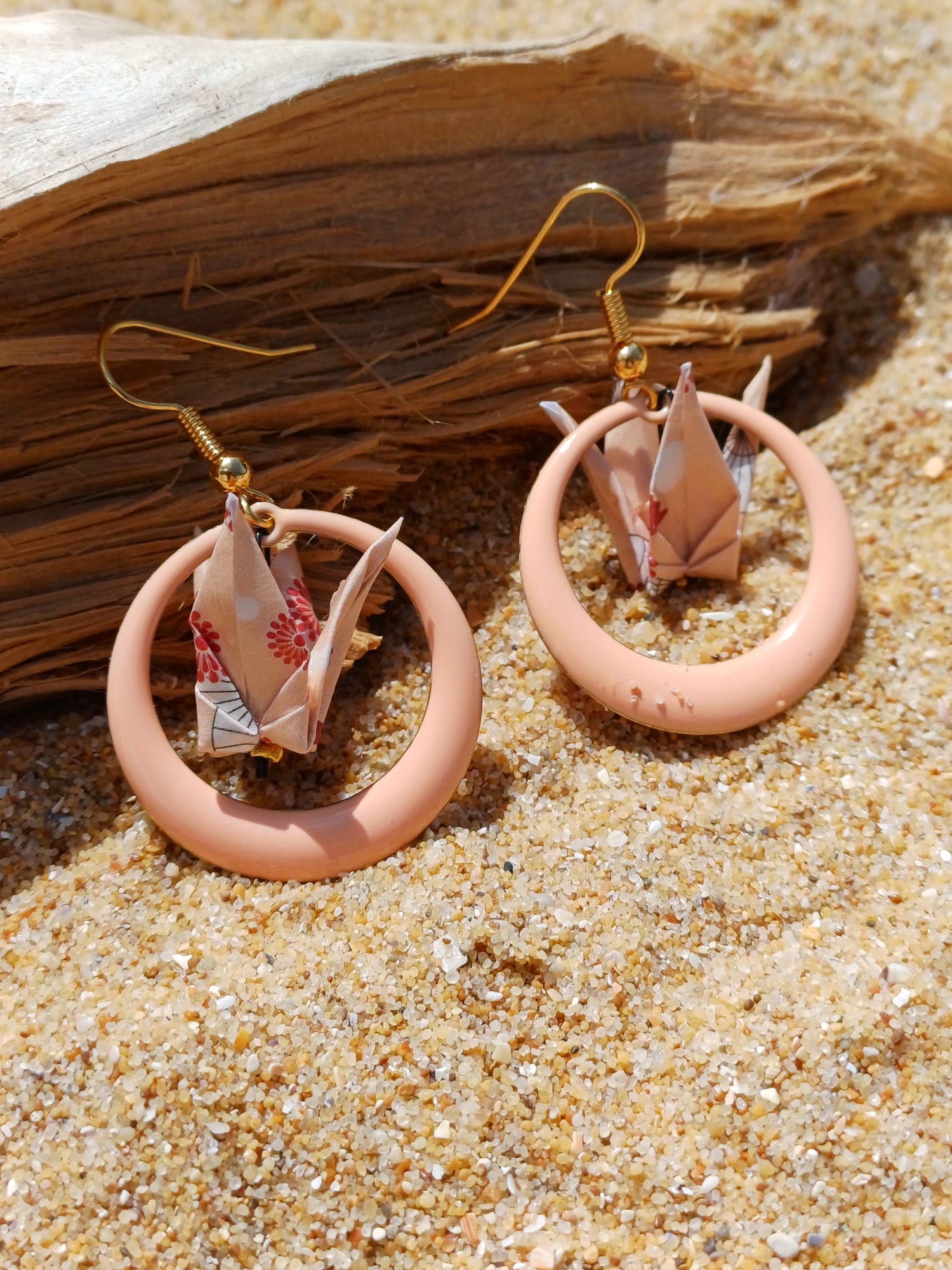 Boucles d'oreilles origami grue beige à motifs floraux dans créole rose poudré, posées sur sable avec bois flotté