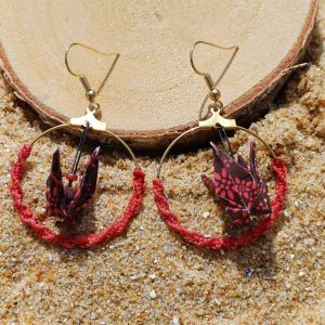 Paire de boucles d'oreilles origami grue bordeaux créole macramé rouge posées contre rondelle de bois dans le sable