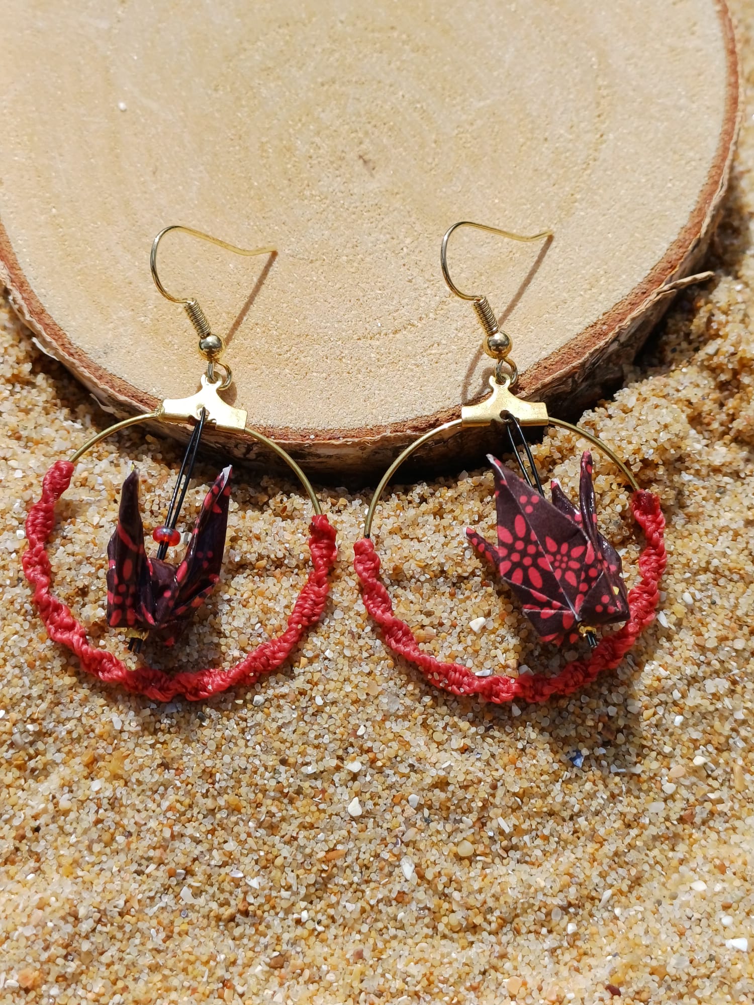 Paire de boucles d'oreilles origami grue bordeaux créole macramé rouge posées contre rondelle de bois dans le sable