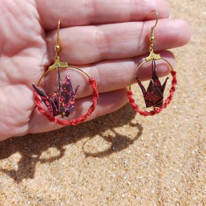 Paire de boucles d'oreilles origami grue bordeaux créole macramé rouge tenues dans une main ouverte, fond sable