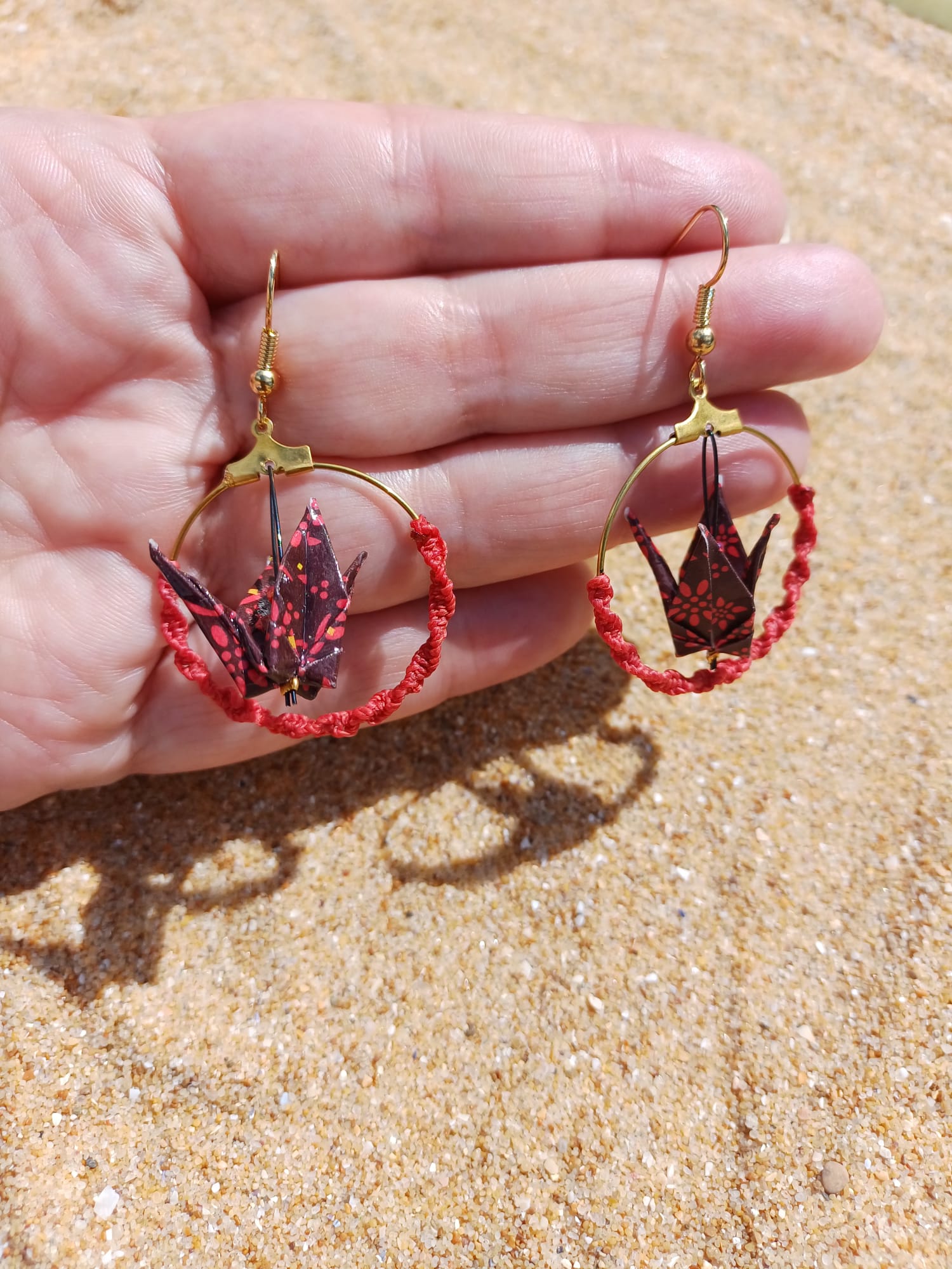 Paire de boucles d'oreilles origami grue bordeaux créole macramé rouge tenues dans une main ouverte, fond sable