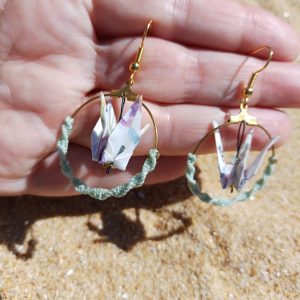 Paire de boucles d'oreilles origami grue blanche créole macramé vert d'eau tenues dans une main ouverte, fond sable