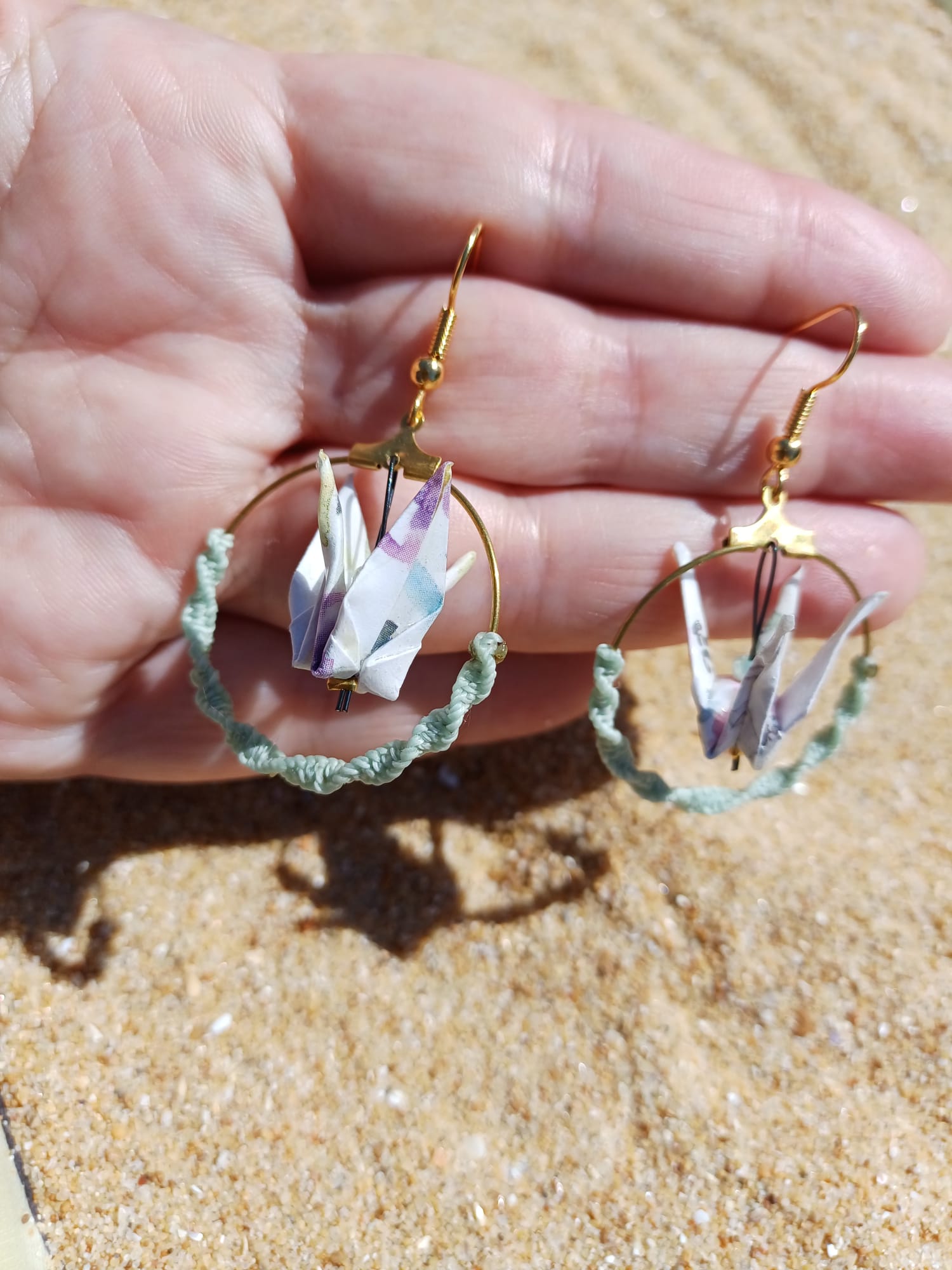 Paire de boucles d'oreilles origami grue blanche créole macramé vert d'eau tenues dans une main ouverte, fond sable