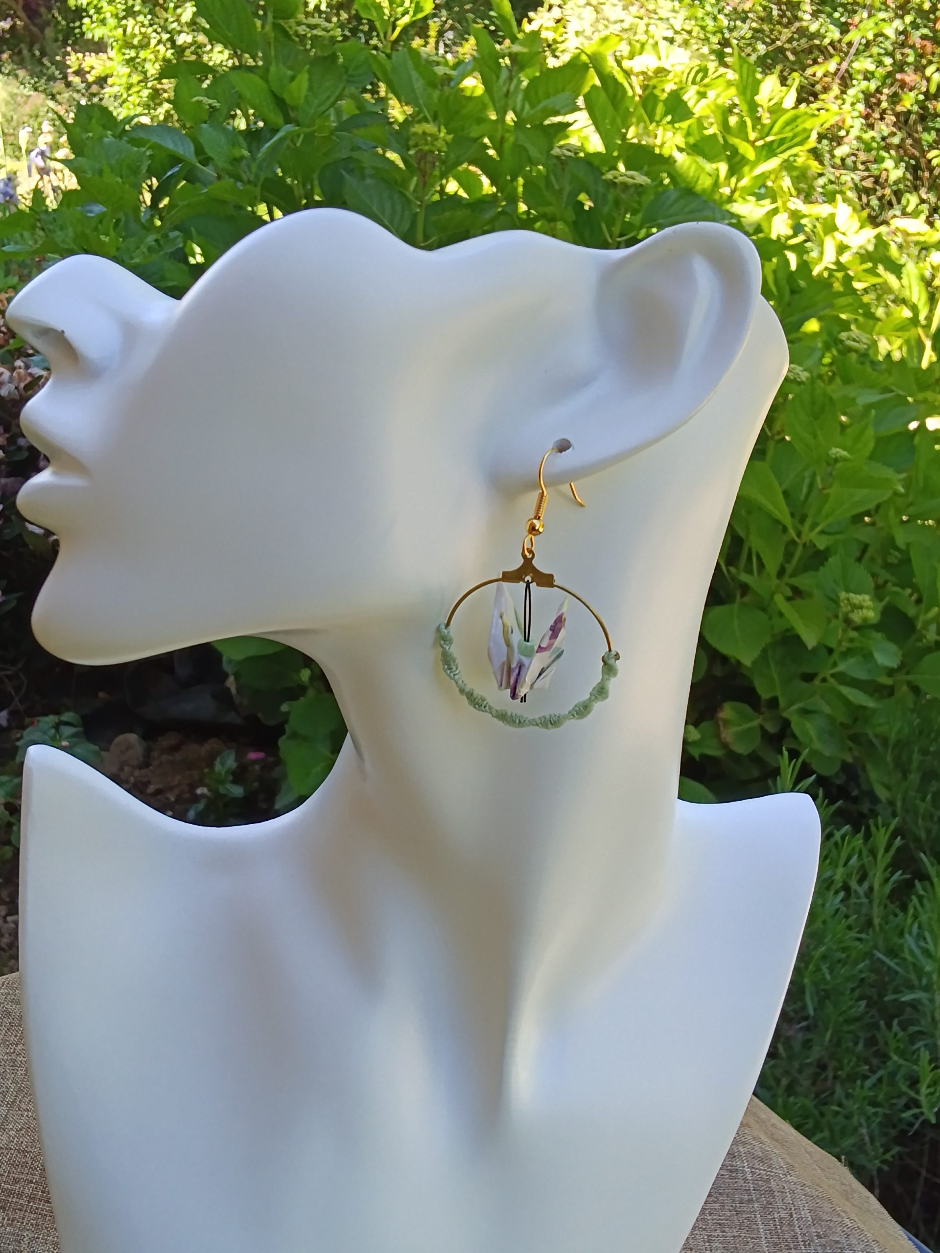 Boucles d'oreilles origami grue blanche aux reflets mauves dans créole macramé vert d'eau, portées sur mannequin bijou fond jardin