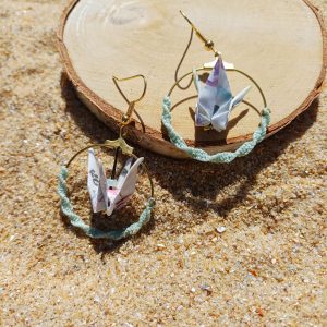Paire de boucles d'oreilles origami grue blanche créole macramé vert d'eau posées sur rondelle de bois dans le sable
