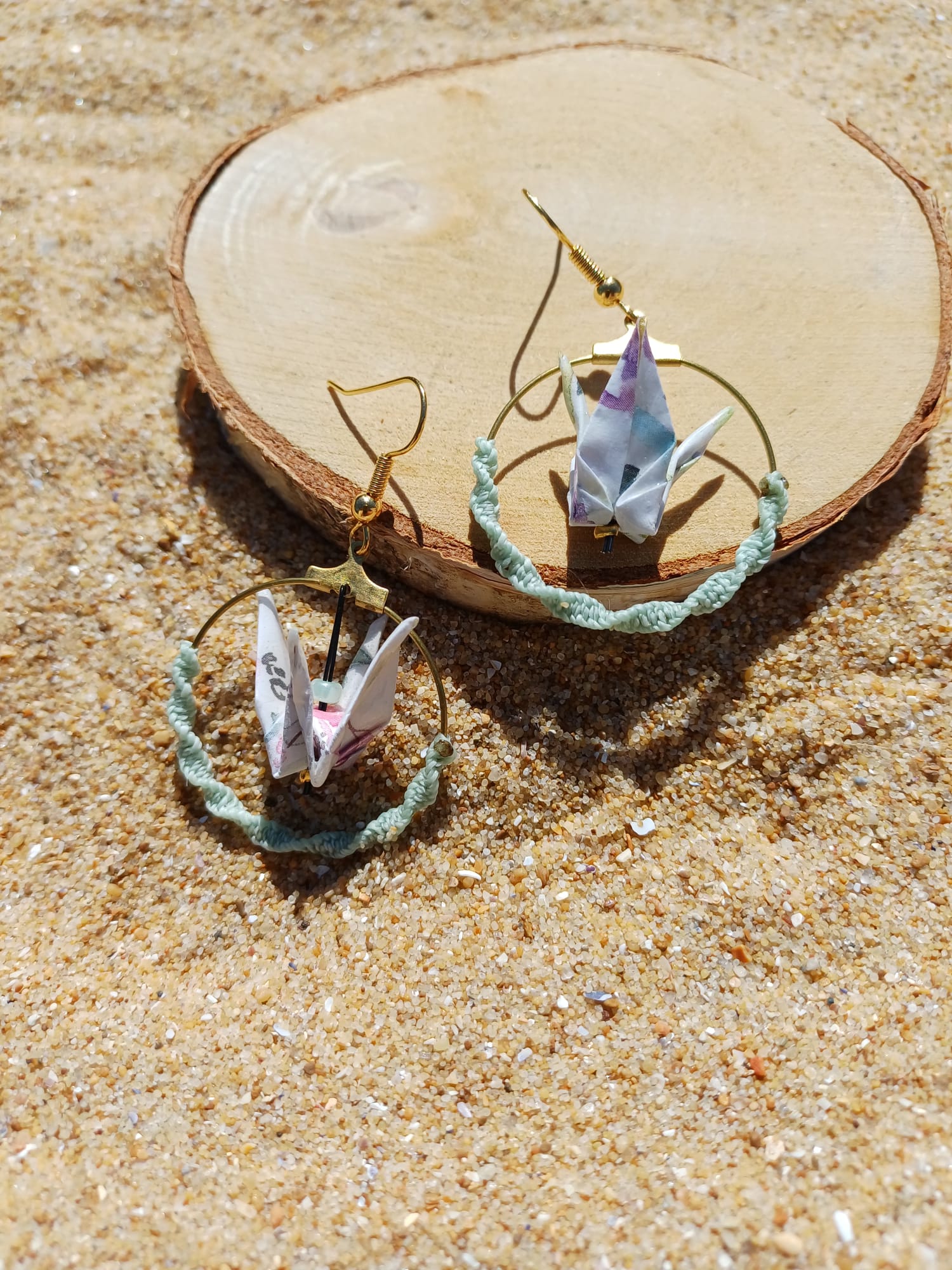 Paire de boucles d'oreilles origami grue blanche créole macramé vert d'eau posées sur rondelle de bois dans le sable
