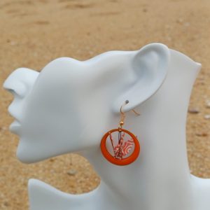 Boucle d'oreille origami grue corail dans un anneau résine orange, portée sur mannequin bijou, fond de sable