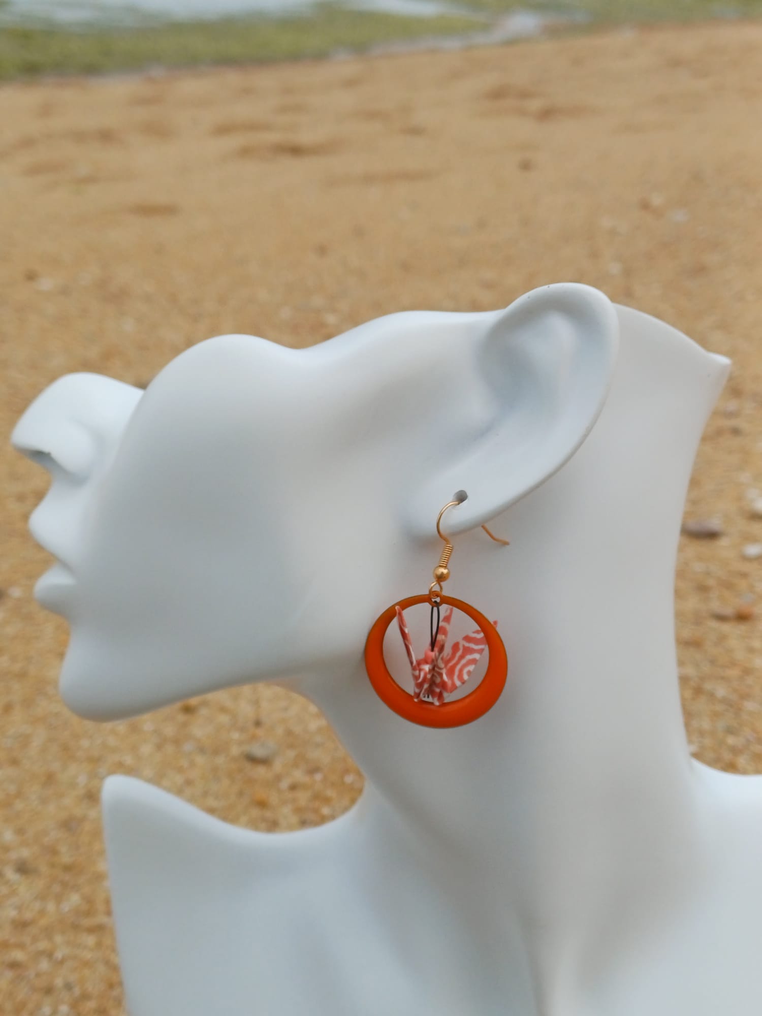Boucle d'oreille origami grue corail dans un anneau résine orange, portée sur mannequin bijou, fond de sable