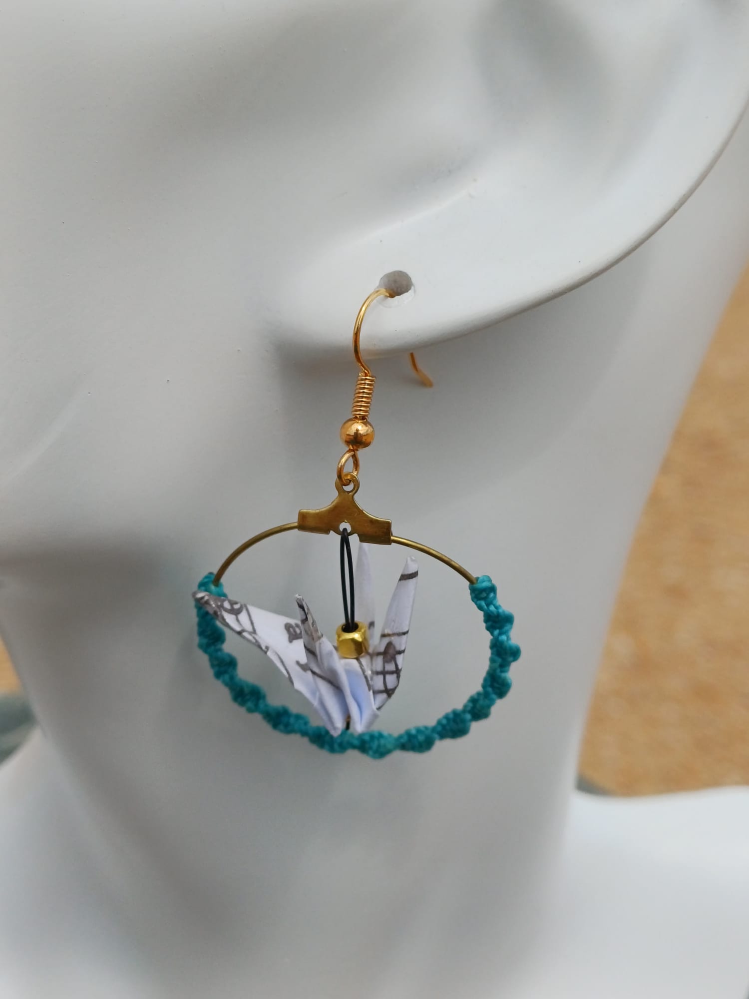 Gros plan sur une boucle d'oreille origami grue en papier à musique, cerceau doré et cordon turquoise