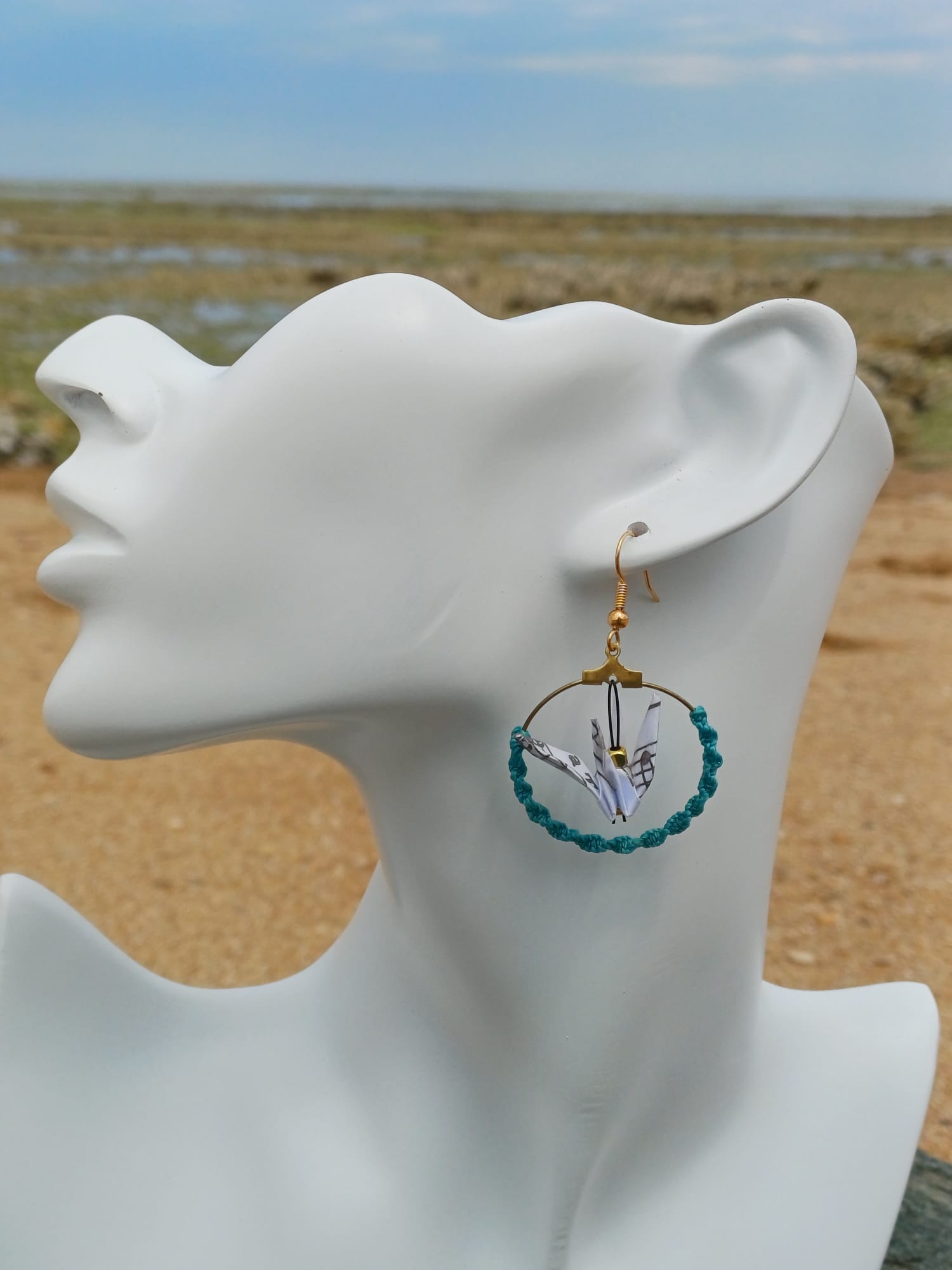 Boucle d'oreille origami grue lagon, cerceau doré et cordon turquoise tressé, portée sur mannequin bijou, fond de marais et sable