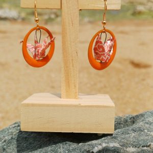 Paire de boucles d'oreilles origami grues corail dans des anneaux résine orange, suspendues sur présentoir bois, fond de marais et sable