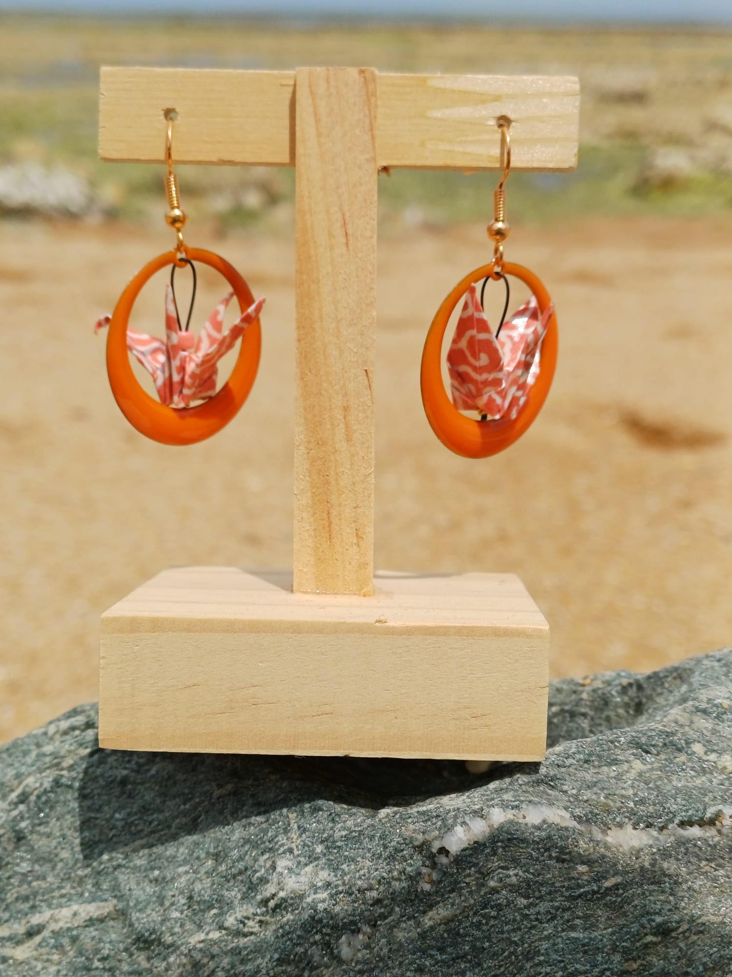 Paire de boucles d'oreilles origami grues corail dans des anneaux résine orange, suspendues sur présentoir bois, fond de marais et sable