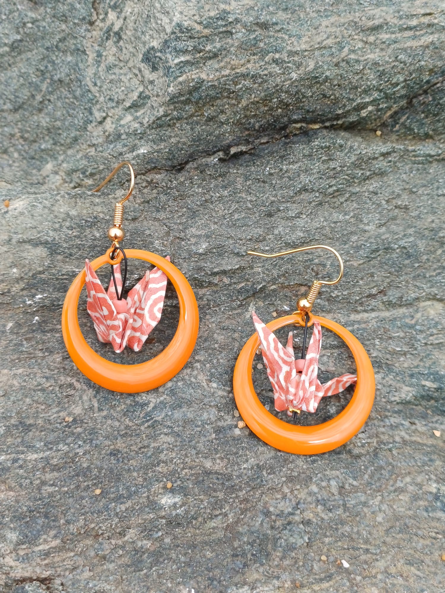 Paire de boucles d'oreilles origami grues corail à motif ondulé dans des anneaux résine orange, posées sur une pierre