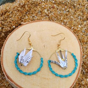 Paire de boucles d'oreilles origami grues en papier à musique dans des cerceaux dorés et cordon turquoise, posées sur un rondin de bois, fond de sable