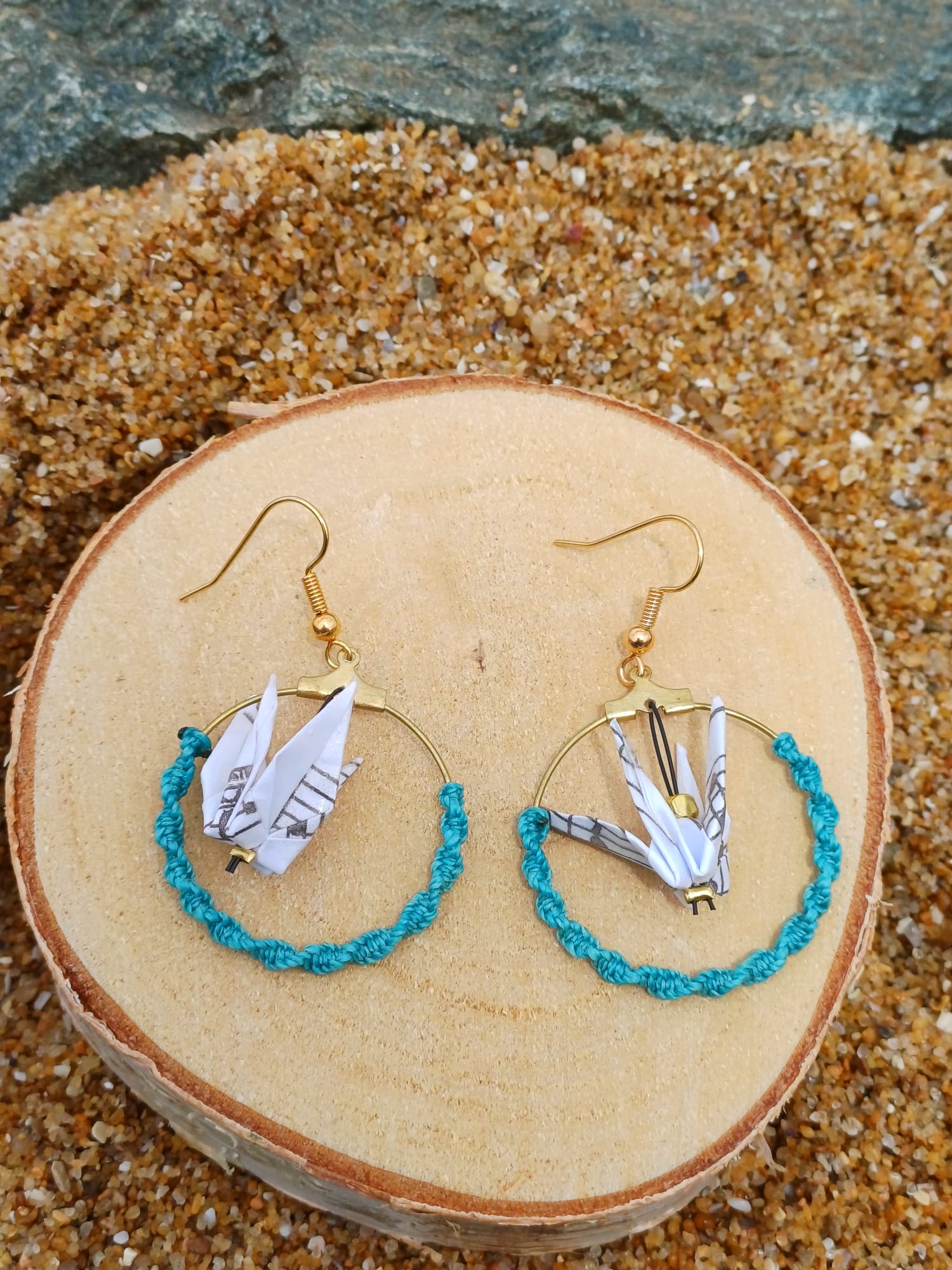 Paire de boucles d'oreilles origami grues en papier à musique dans des cerceaux dorés et cordon turquoise, posées sur un rondin de bois, fond de sable