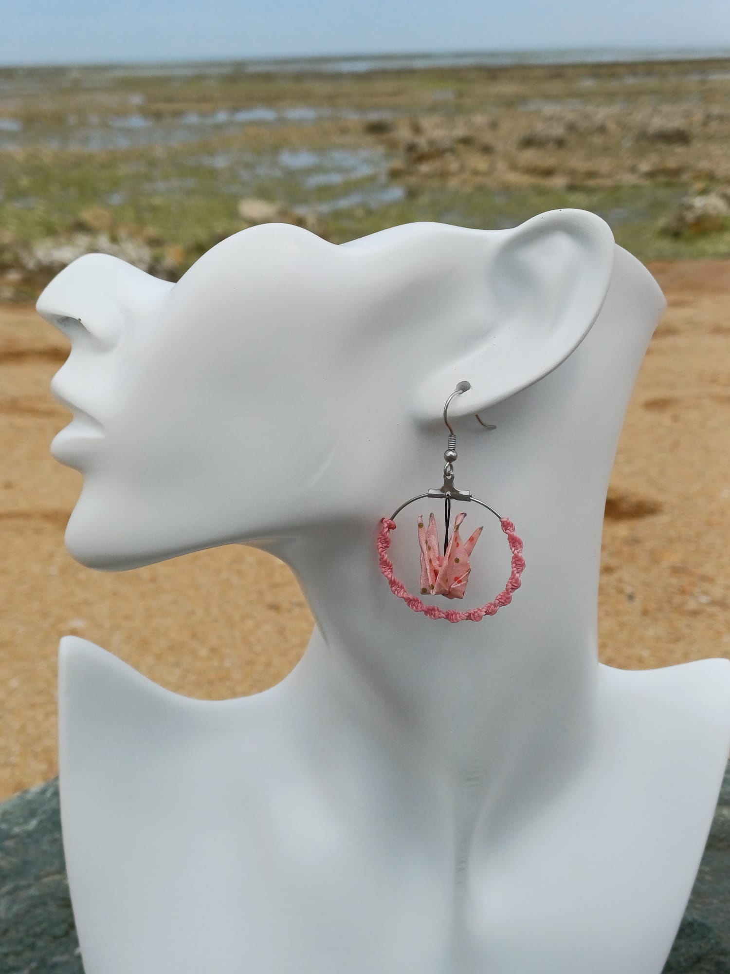 Boucle d'oreille origami grue rose portée sur mannequin bijou, fond de marais et sable
